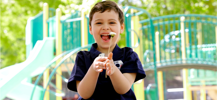 A child in a blue t-shirt stands in front of a teal jungle gym and smiles at the camera while holding a dandelion.
