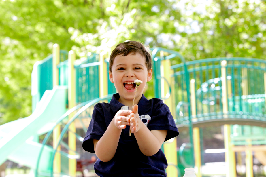 A child stands in front of a jungle gym and smiles while holding a dandelion.