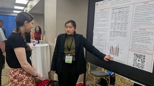 A COMPASS student stands in front of a poster at a conference and presents statistics to a patron.