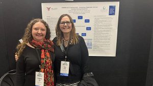 Dr. Lindsey Ogle and Dr. Lisa Ruble smile at the camera in front of a poster board they were presenting at a conference